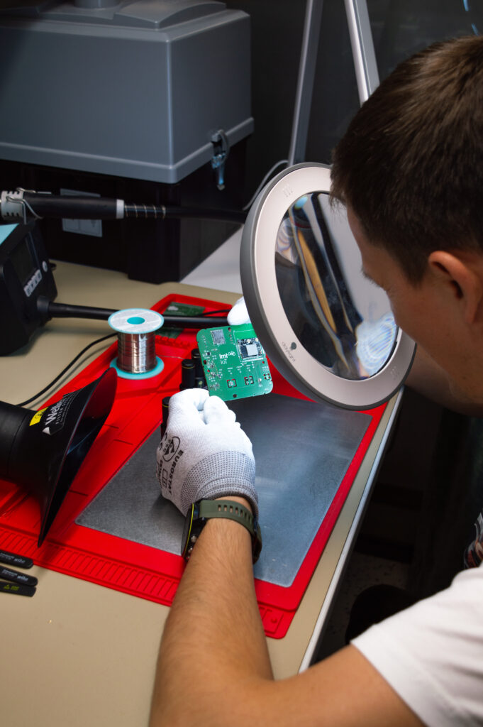 Technician inspecting an LMT IoT circuit board under a magnifying lamp