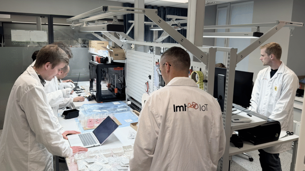 LMT IoT engineers working in a hardware lab, assembling and testing electronic components at multiple workstations with laptops, tools, and prototyping equipment.