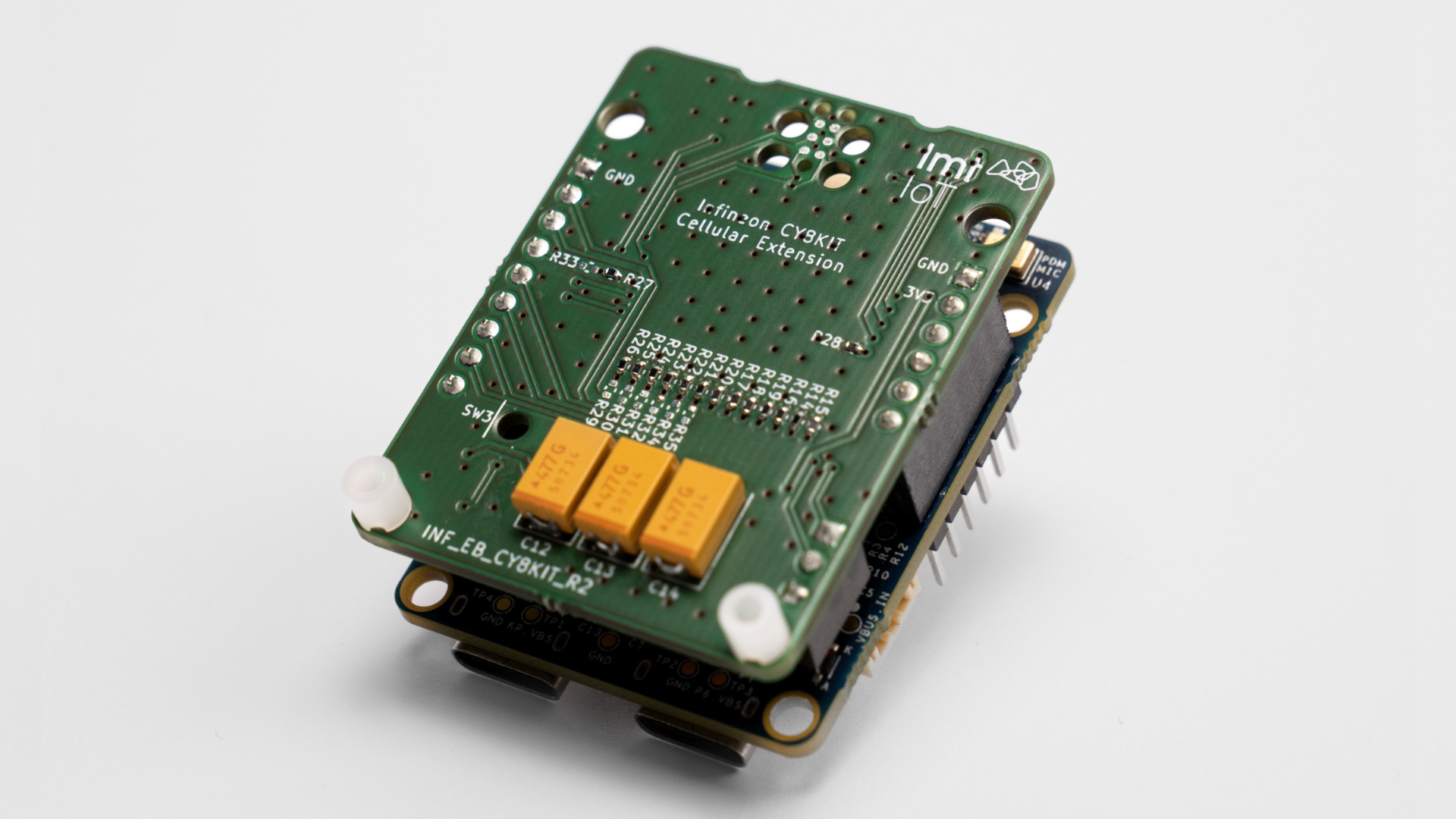 A detailed close-up photo of the LMT IoT Infineon CY8KIT Cellular Extension board attached to an Infineon hardware development kit. The compact green PCB features electronic components, connectors, and the LMT IoT logo. The hardware is designed to simplify LTE-M and NB-IoT connectivity for edge AI and low-power IoT product development.
