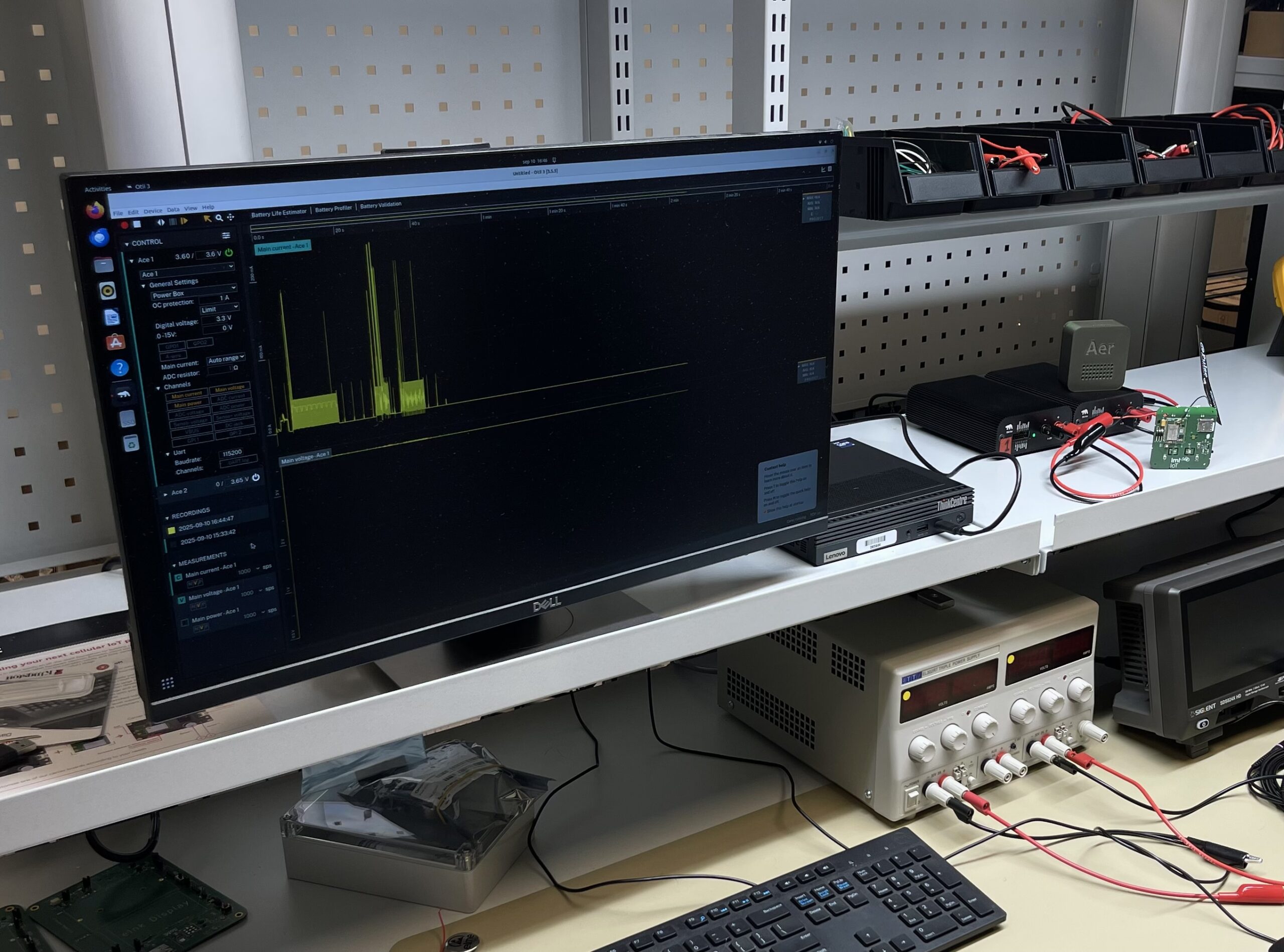 A computer monitor on a workbench displays a power consumption graph with yellow spikes alongside a power supply and testing equipment in the LMT IoT lab